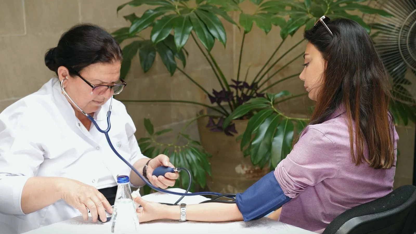 A clinician in a white coat taking a seated woman's blood pressure with a cuff and stethoscope