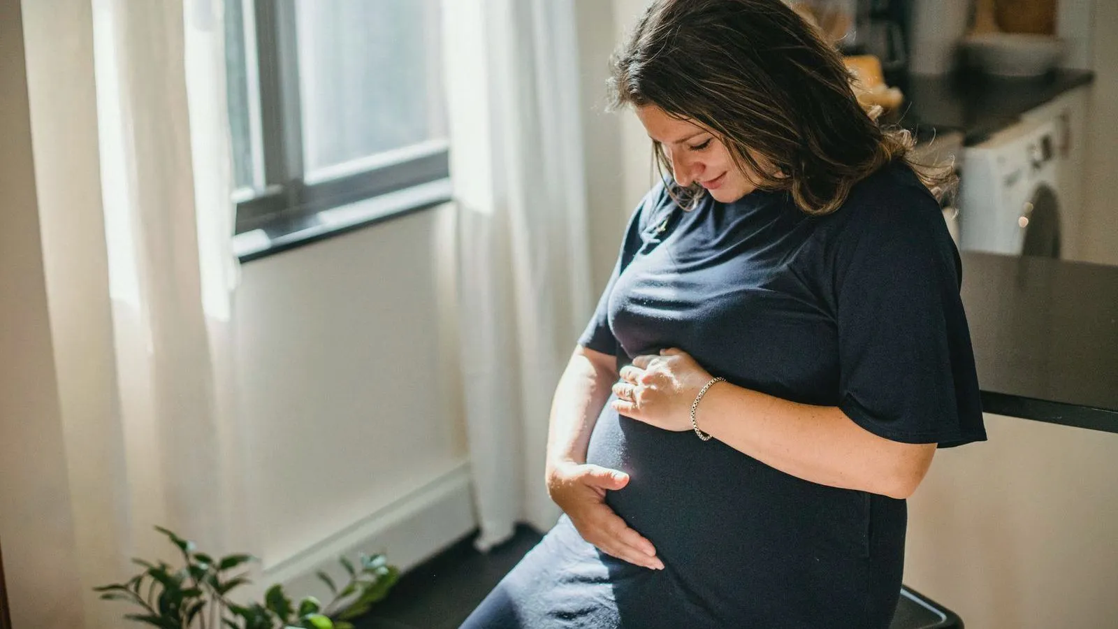 A pregnant woman standing by a kitchen window with a hand on her bump