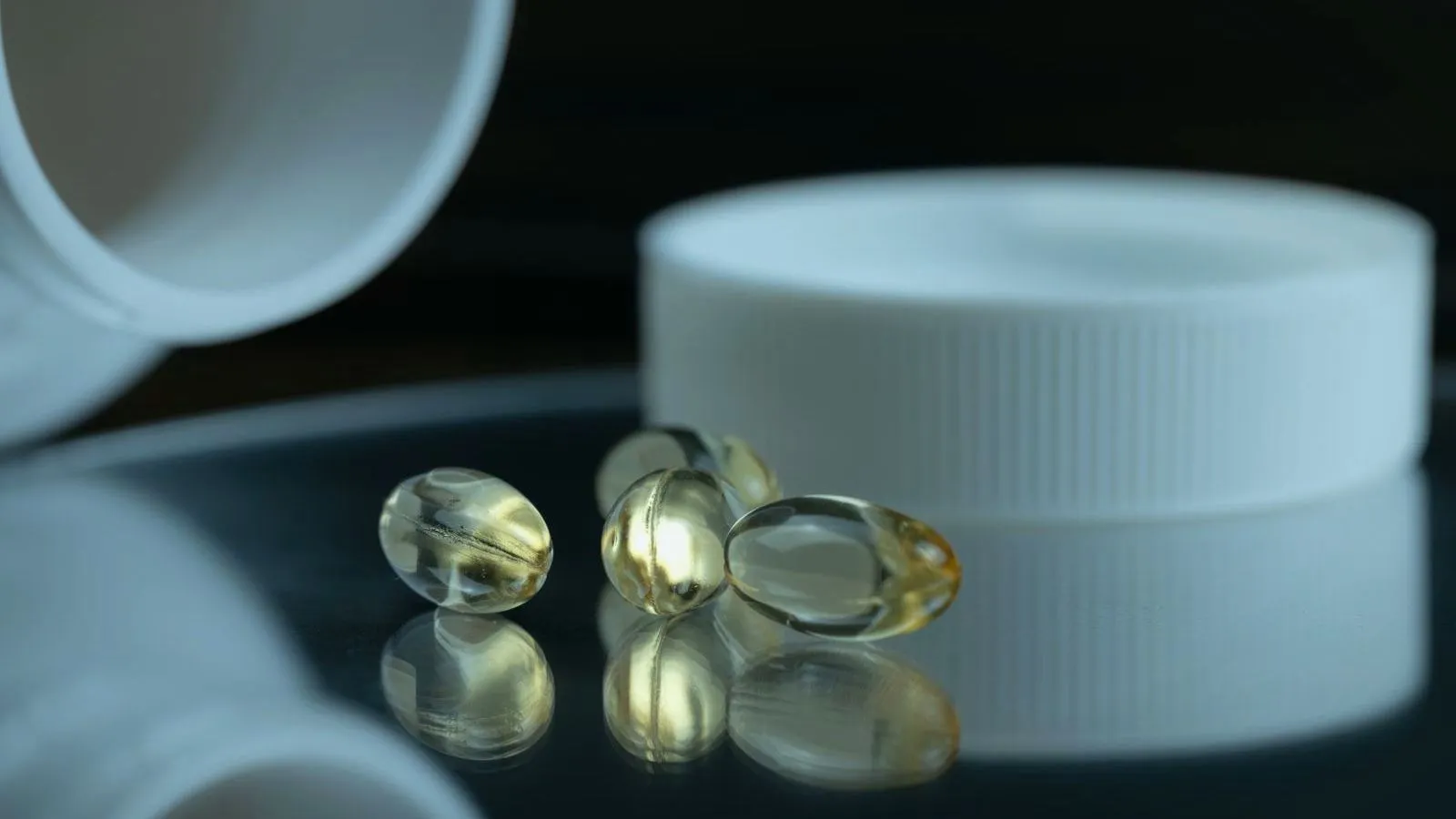 Three clear yellow softgel capsules lie on a dark reflective surface beside a white bottle and its cap.