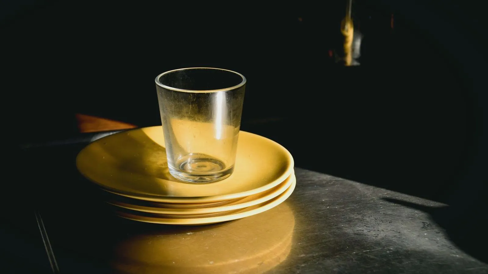 An empty drinking glass standing on a stack of yellow plates on a dark table, lit from one side.
