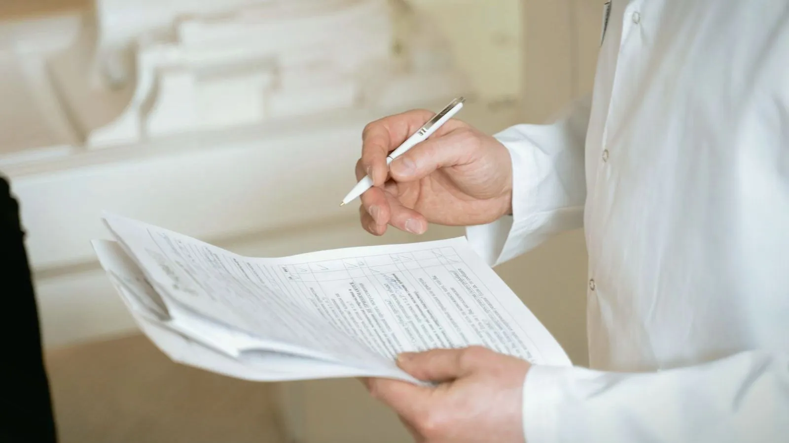 The hands of a person in a white coat hold printed forms and a pen.