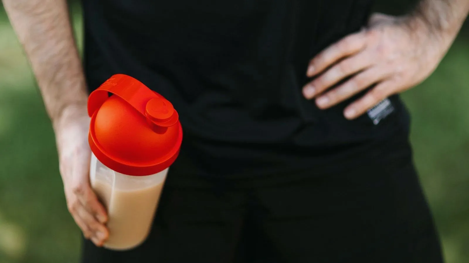 A person in a black shirt holds a shaker bottle with a red lid at the hip, the other hand resting on the waist, outdoors.