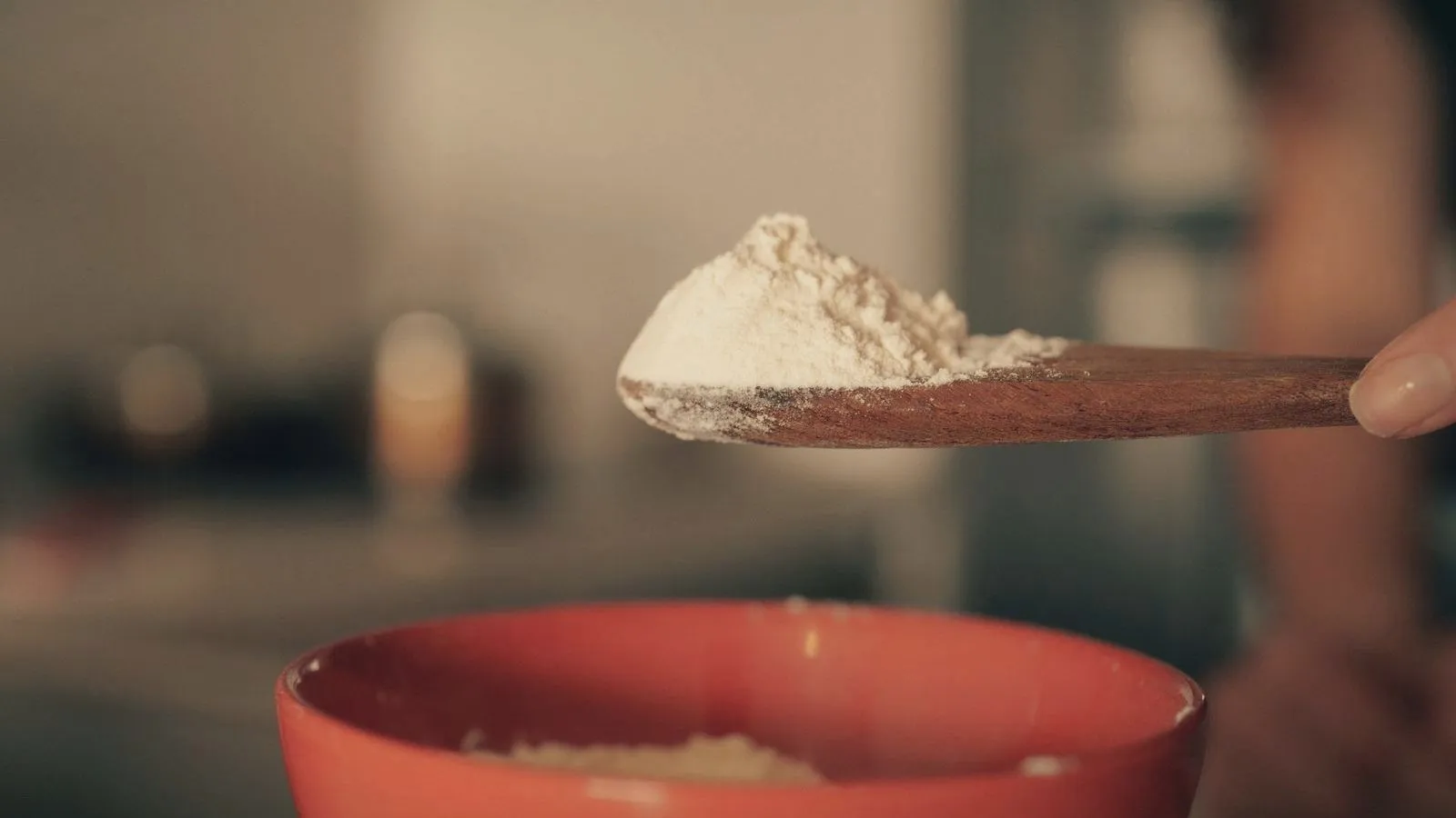 A wooden spoon holding a heaped mound of pale powder above a red bowl