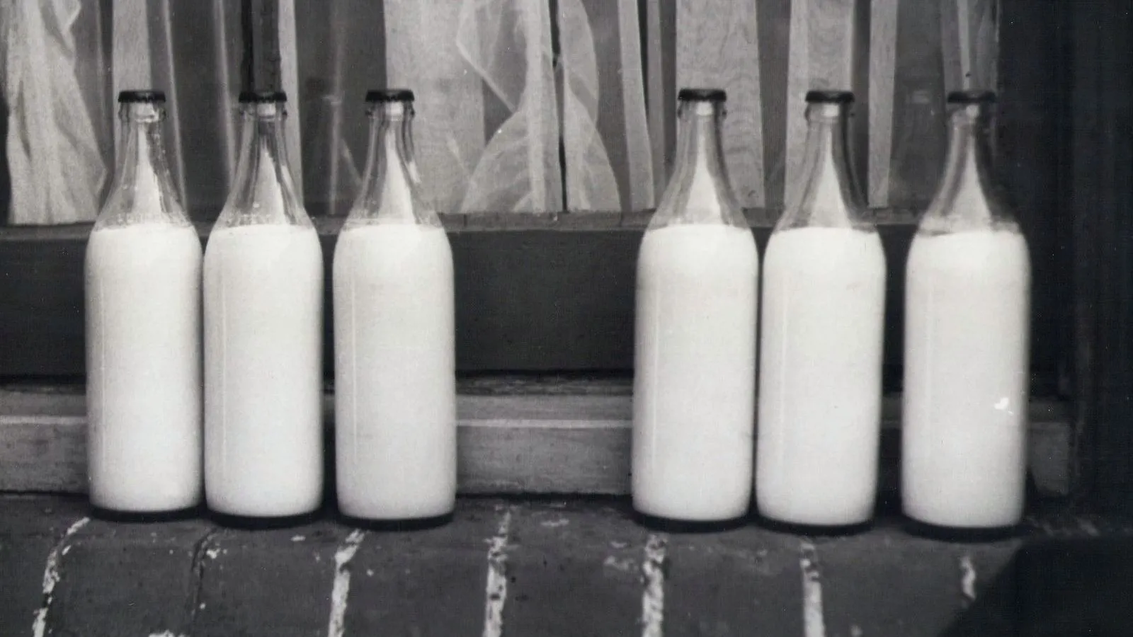 Six unlabelled glass bottles of milk stand in two groups of three on a brick windowsill, in black and white.