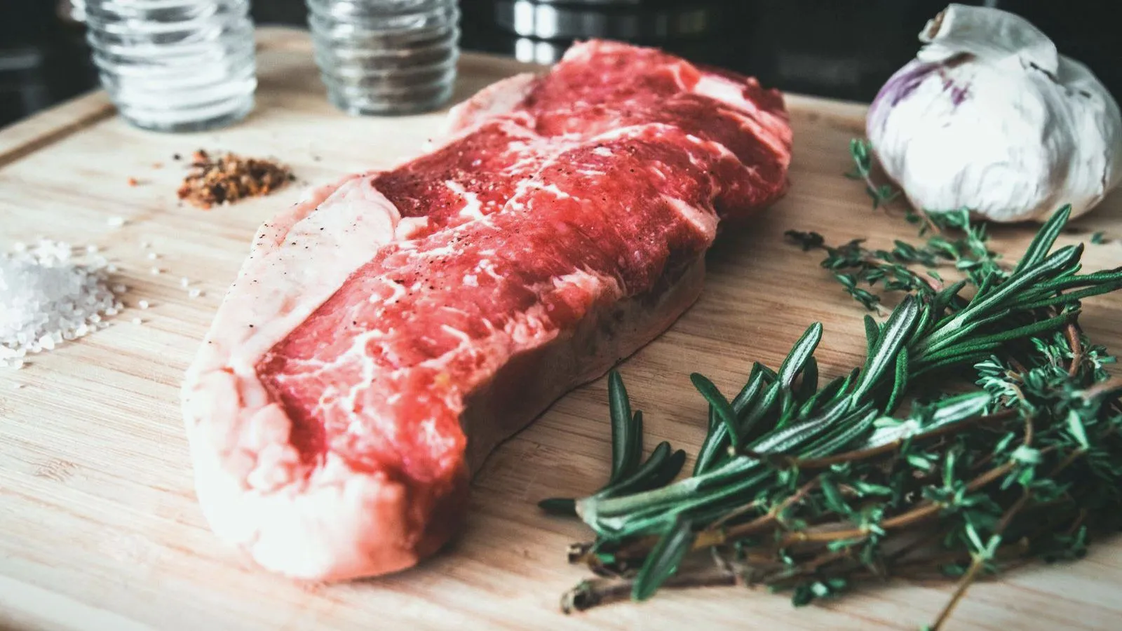 A raw steak on a wooden board beside rosemary, a bulb of garlic and coarse salt.