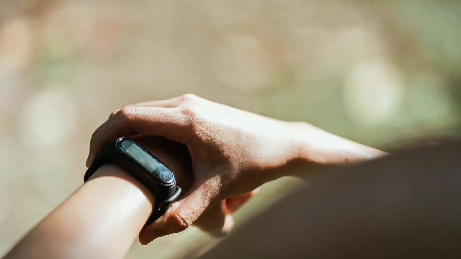 A wrist wearing a plain black activity band, with the other hand touching it, against a blurred outdoor background.