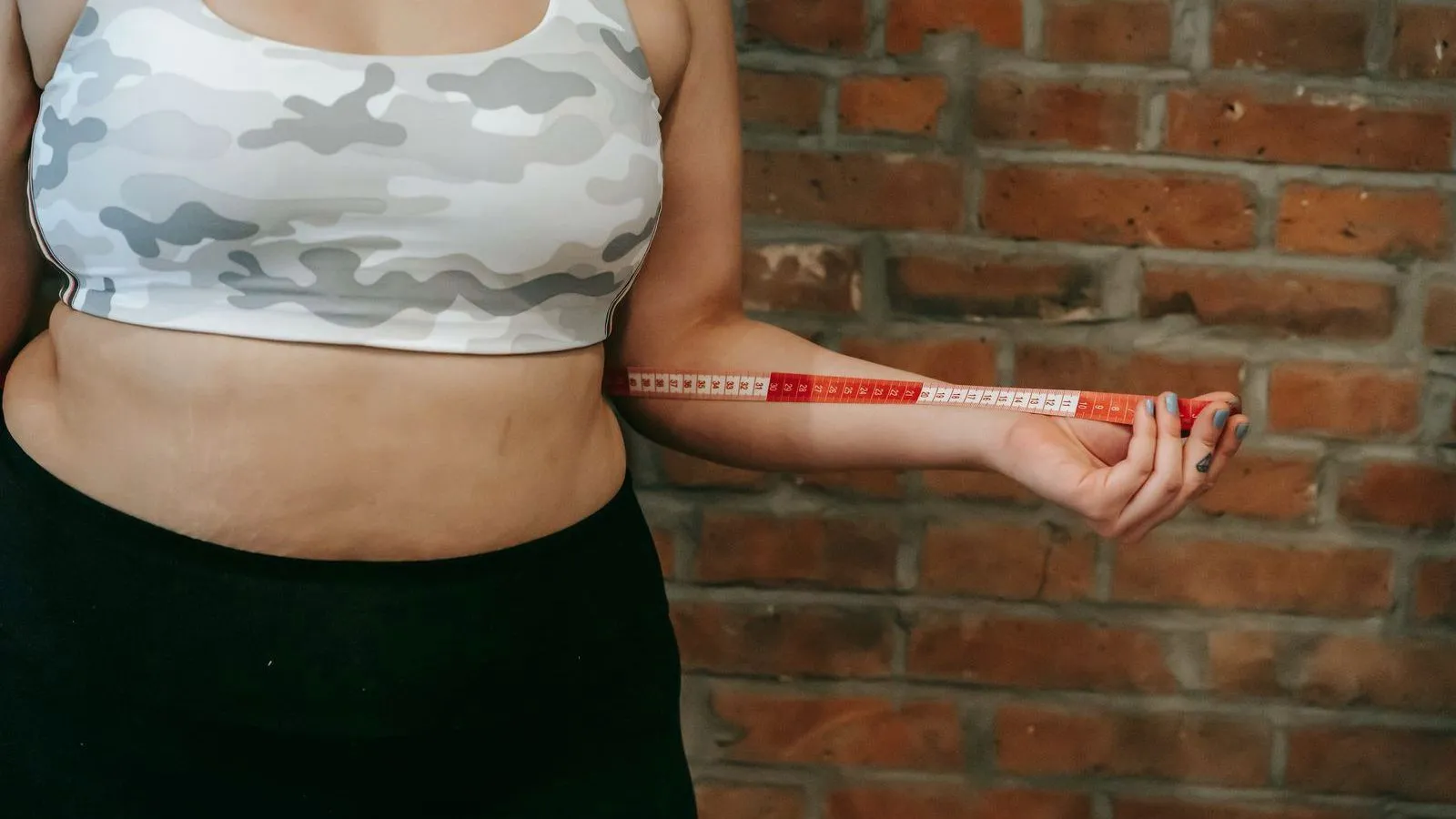 A person in a sports top and dark leggings stands against a brick wall holding a tape measure out from the waist, face out of frame.