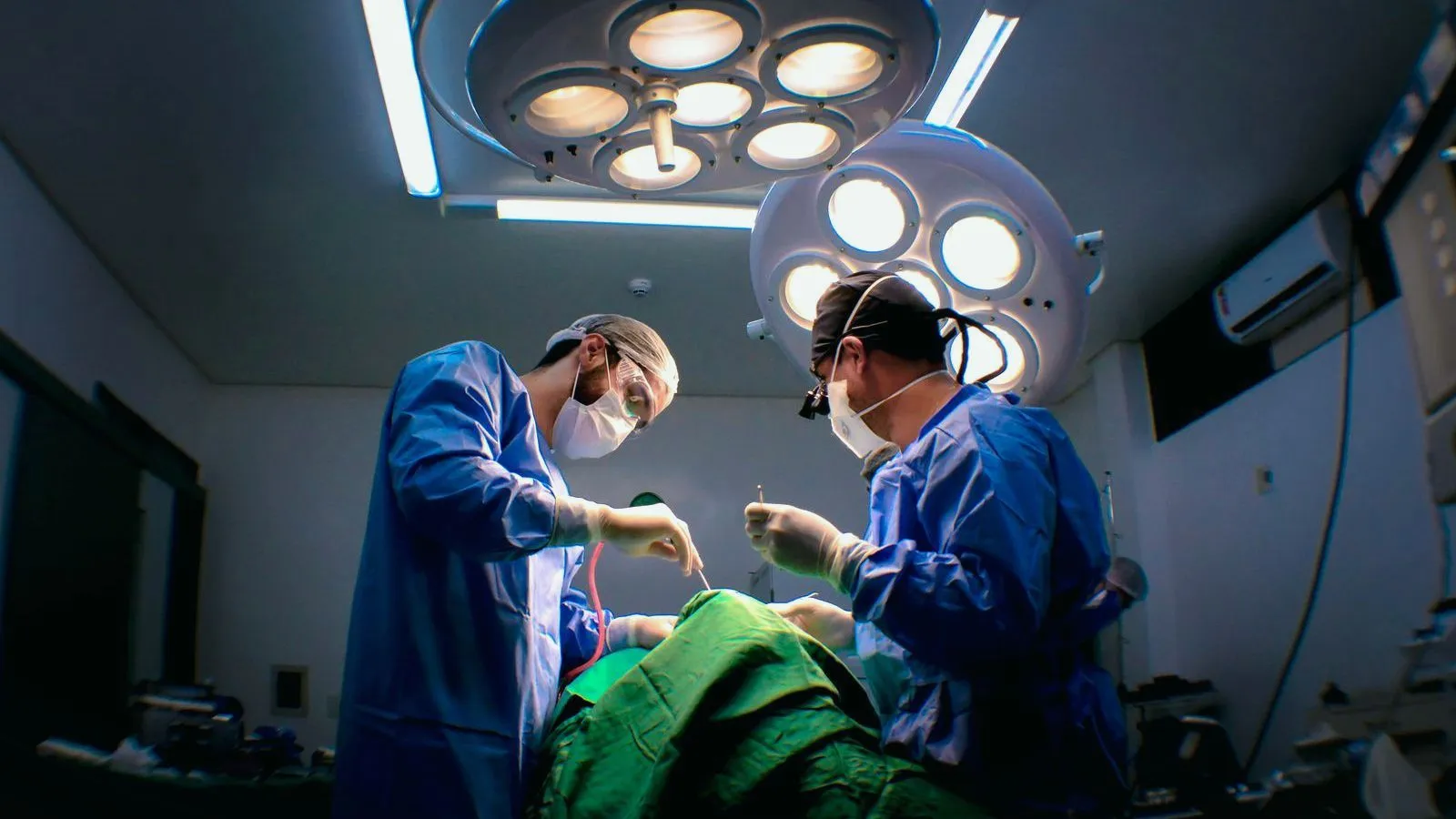 Two surgeons in blue gowns, masks and caps operate on a patient under green drapes beneath large round theatre lights.