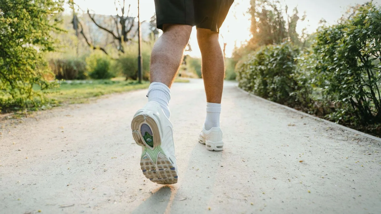 A runner's legs and white shoes on a sunlit park path, seen from behind.