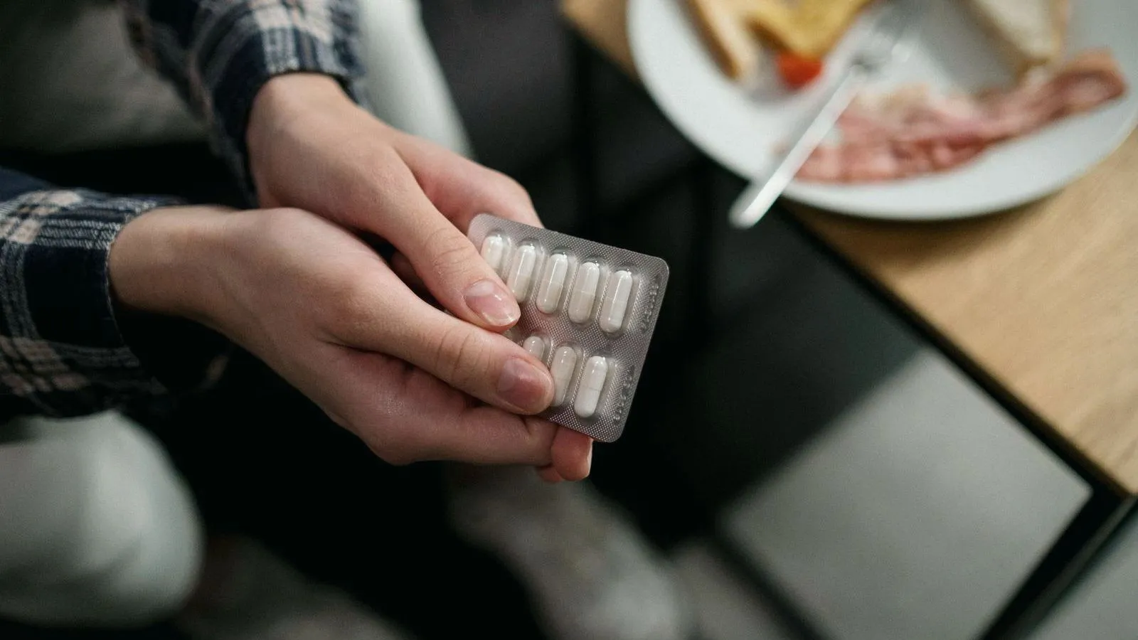 Hands holding a blister pack of white capsules, with a breakfast plate on the table behind.