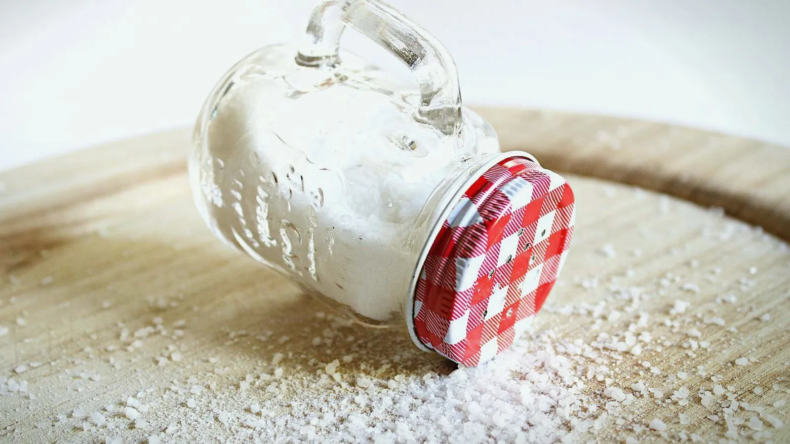 A tipped-over glass salt shaker with a checked lid spilling coarse salt onto a wooden board