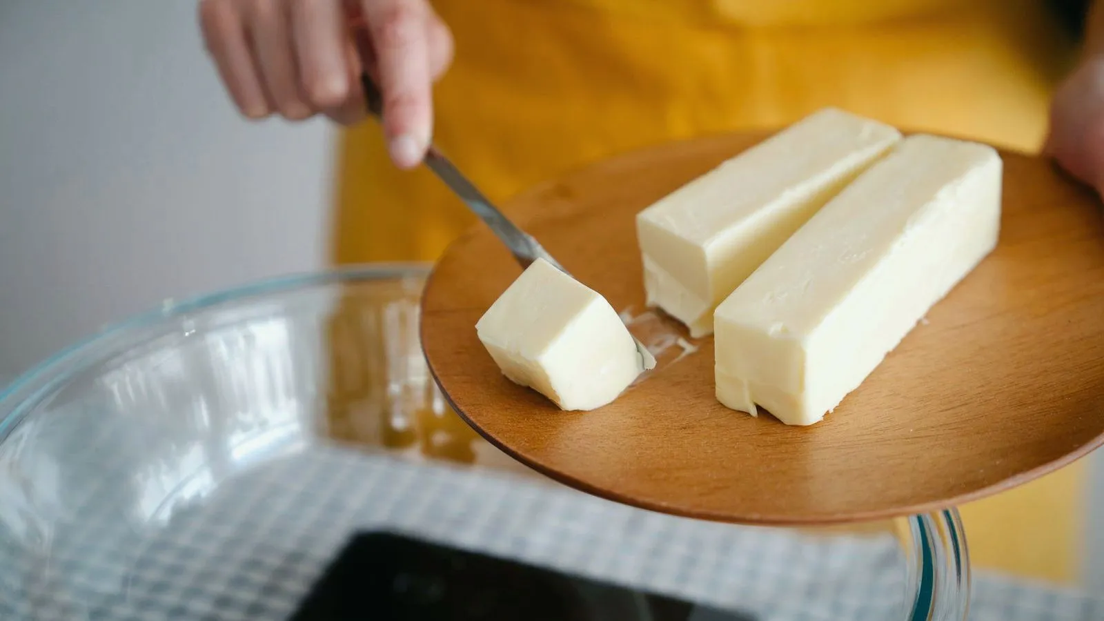 Two sticks of butter on a wooden plate, with a hand cutting a piece from one of them with a knife.
