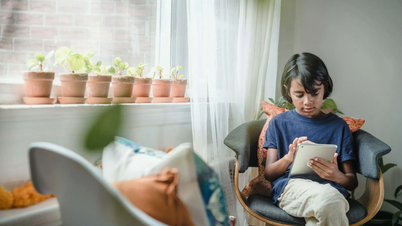 A child in a navy T-shirt sits in an armchair using a tablet, beside a windowsill lined with small potted plants.