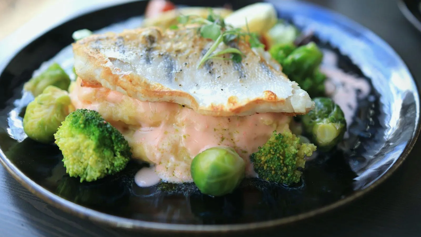 A pan-seared white fish fillet served with broccoli and brussels sprouts on a dark plate