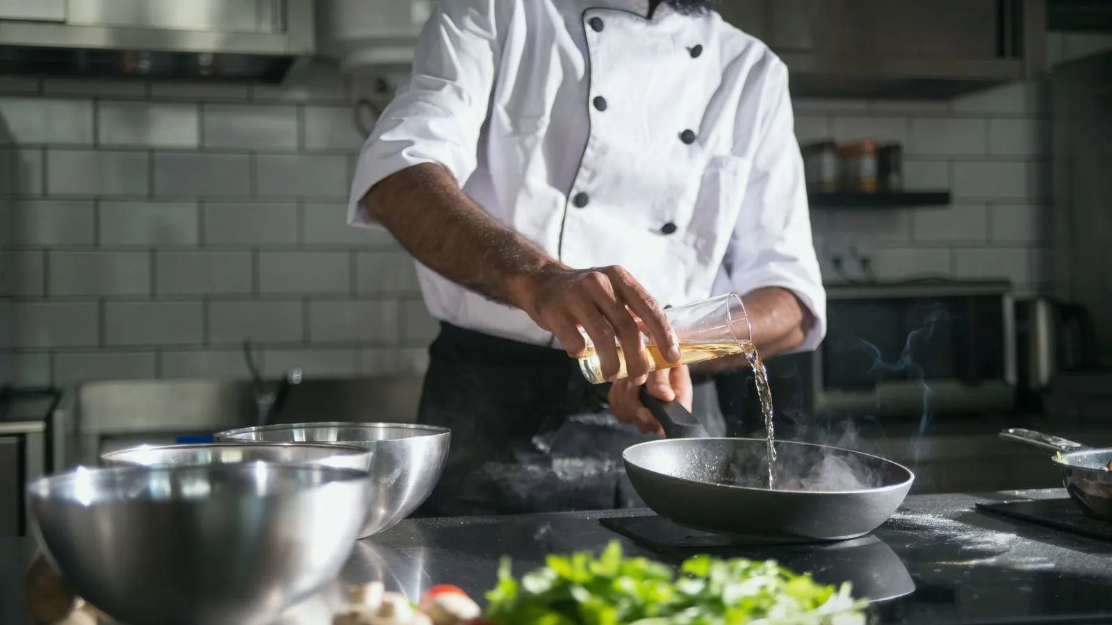 A cook in a white jacket pouring oil from a glass into a hot pan in a kitchen.