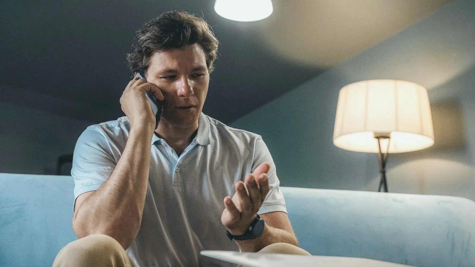 A middle-aged man sitting on a pale sofa at home, holding a phone to his ear and gesturing