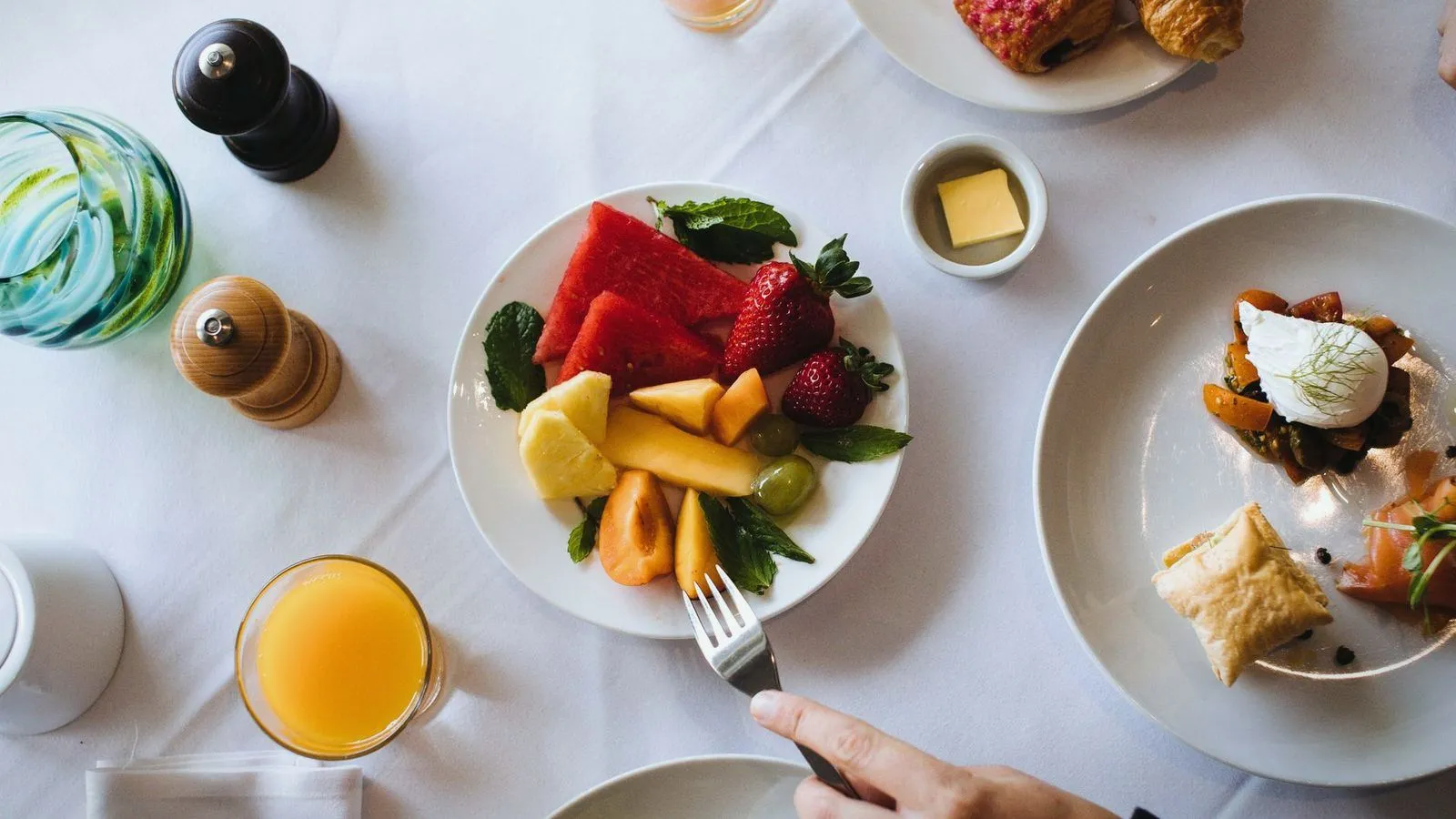 An overhead breakfast table with a plate of fruit and a hand holding a fork
