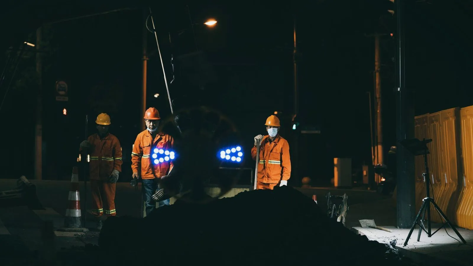 Three road workers in orange high-visibility clothing and hard hats standing at a work site at night.
