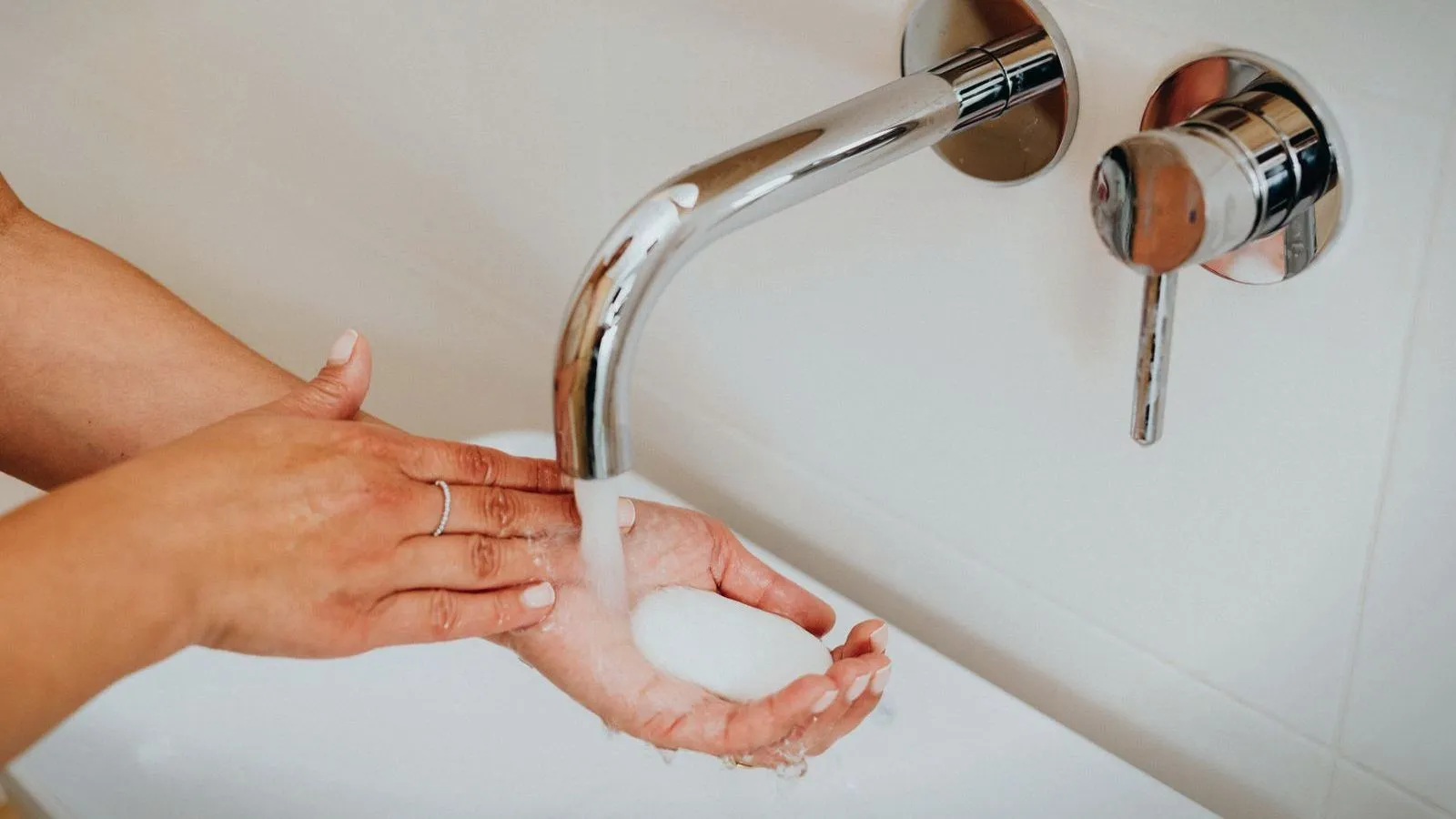 Two hands lathered with soap are held under water running from a chrome wall tap over a white basin.