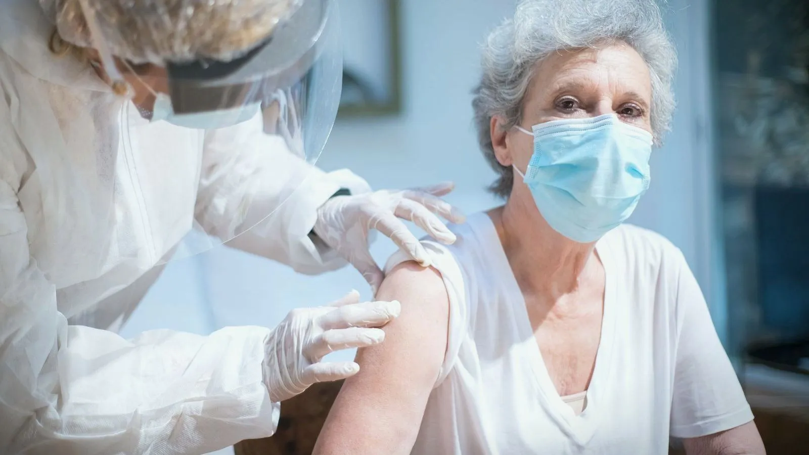 An older person in a face mask receiving an injection from a clinician in protective clothing