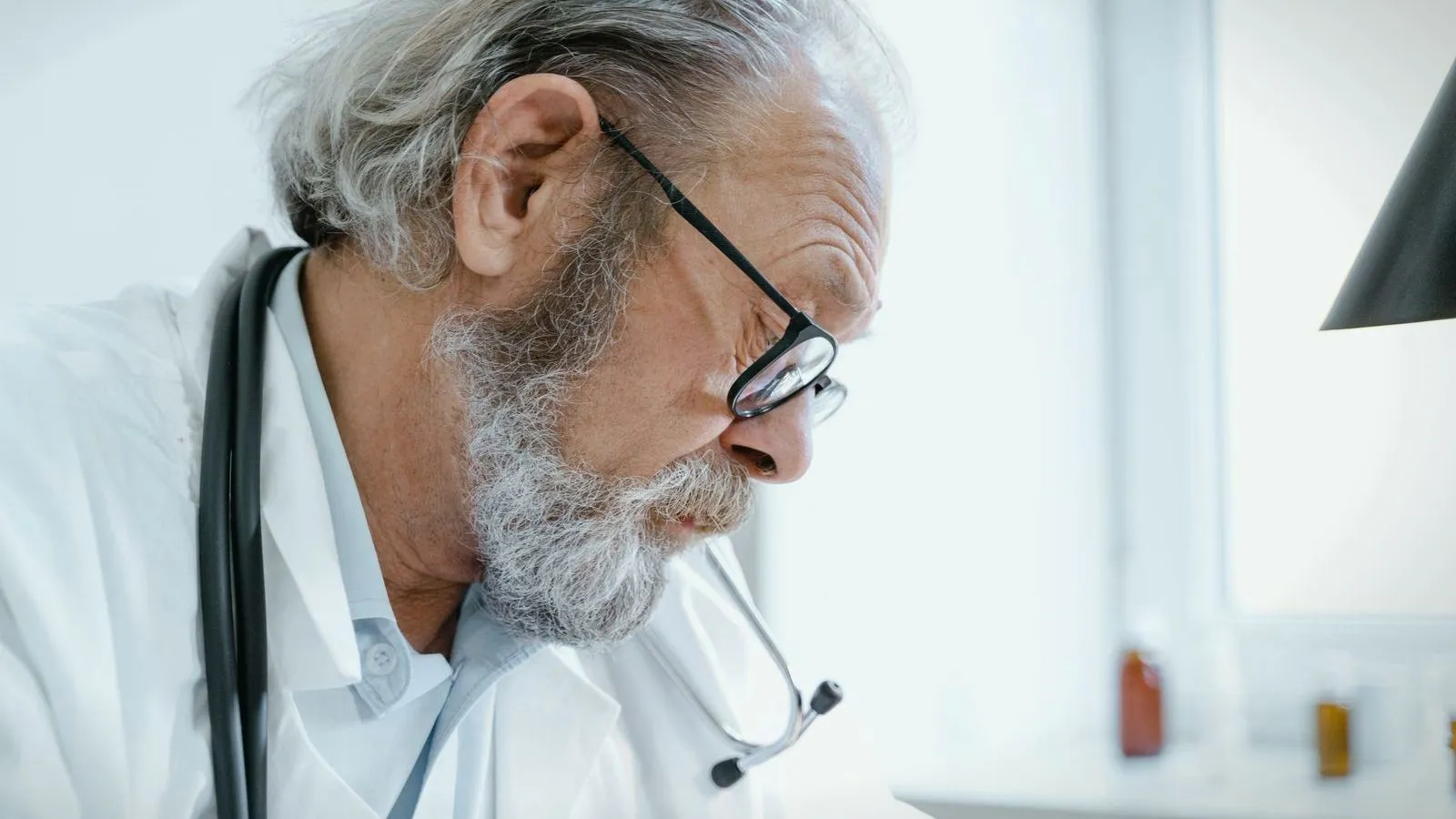 An older doctor with a gray beard and glasses, a stethoscope around the neck, looking down at a desk