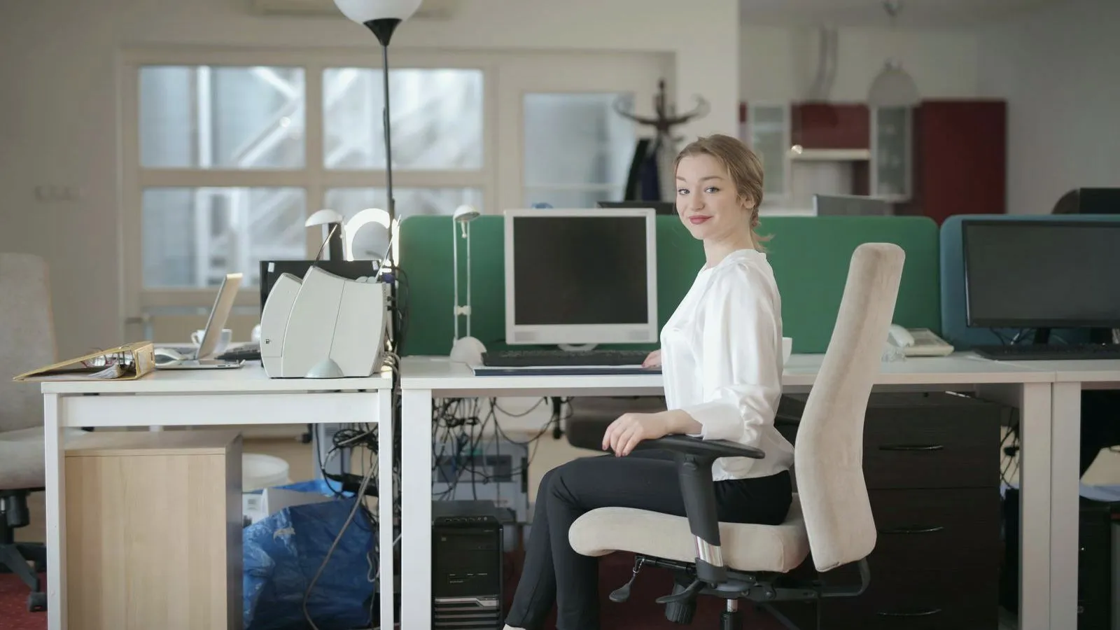 A woman sitting in an office chair at a desk with a computer, looking at the camera.