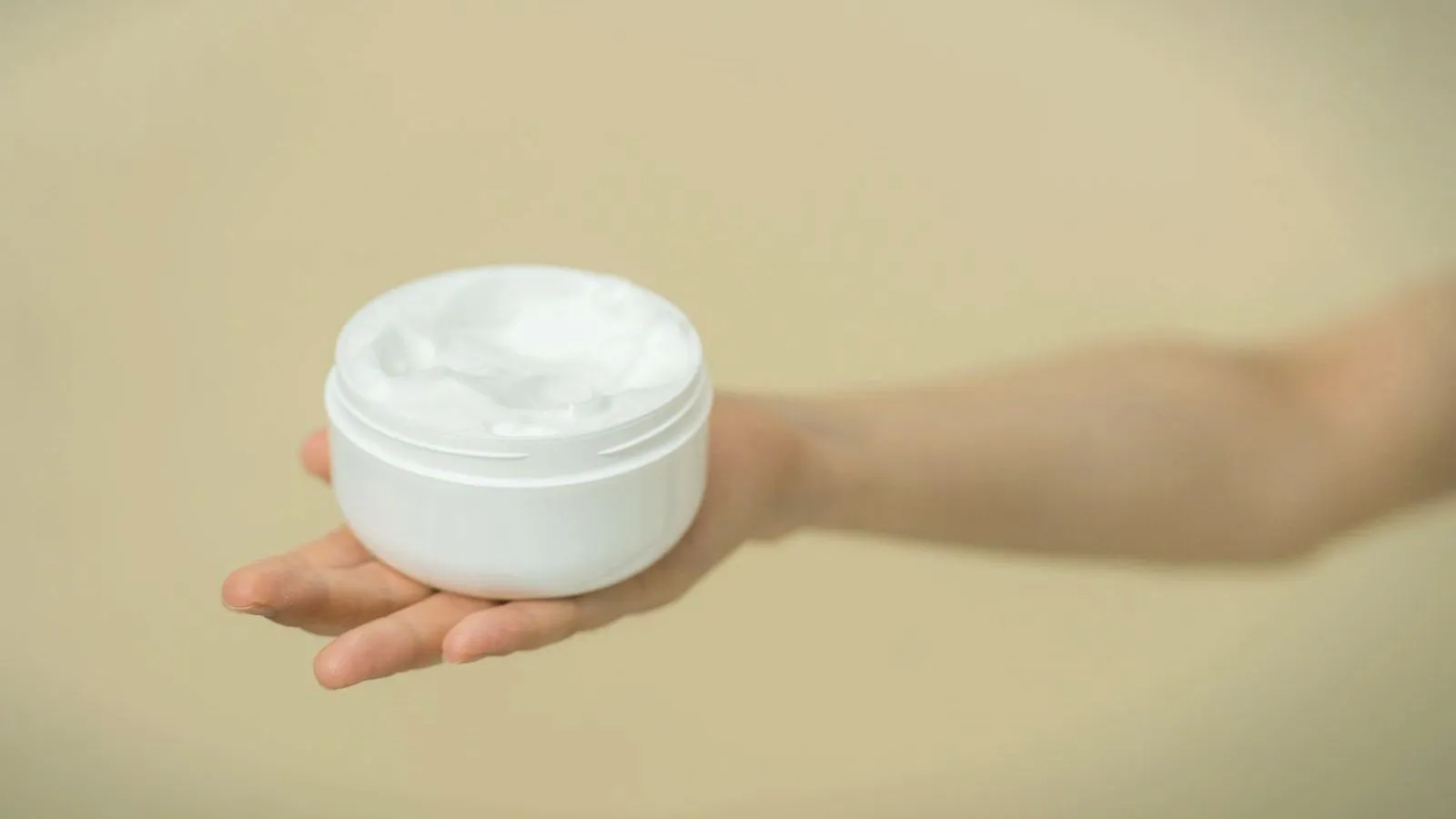 An open, unlabelled white jar of cream sits on an outstretched palm against a beige background.