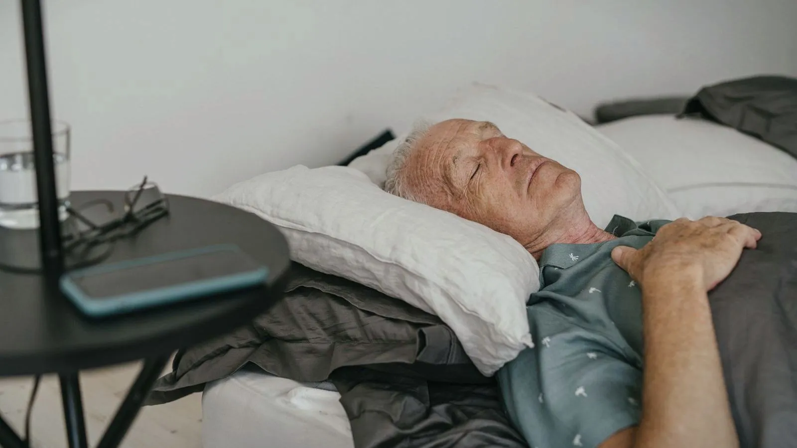 An older man asleep on his back in bed, with his glasses and a phone on the nightstand beside him
