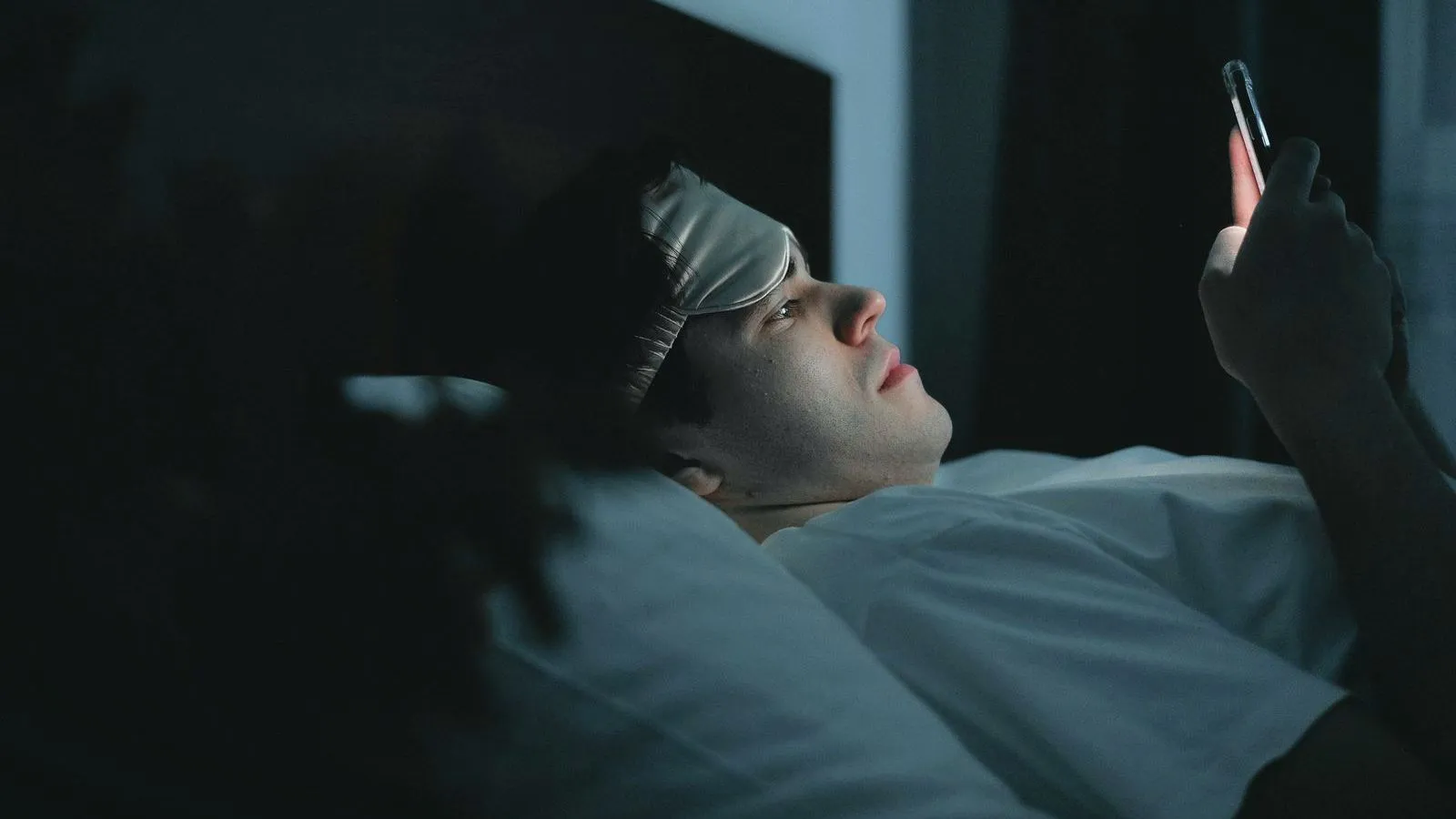 A person lying in bed in the dark with a sleep mask pushed up, looking at a phone