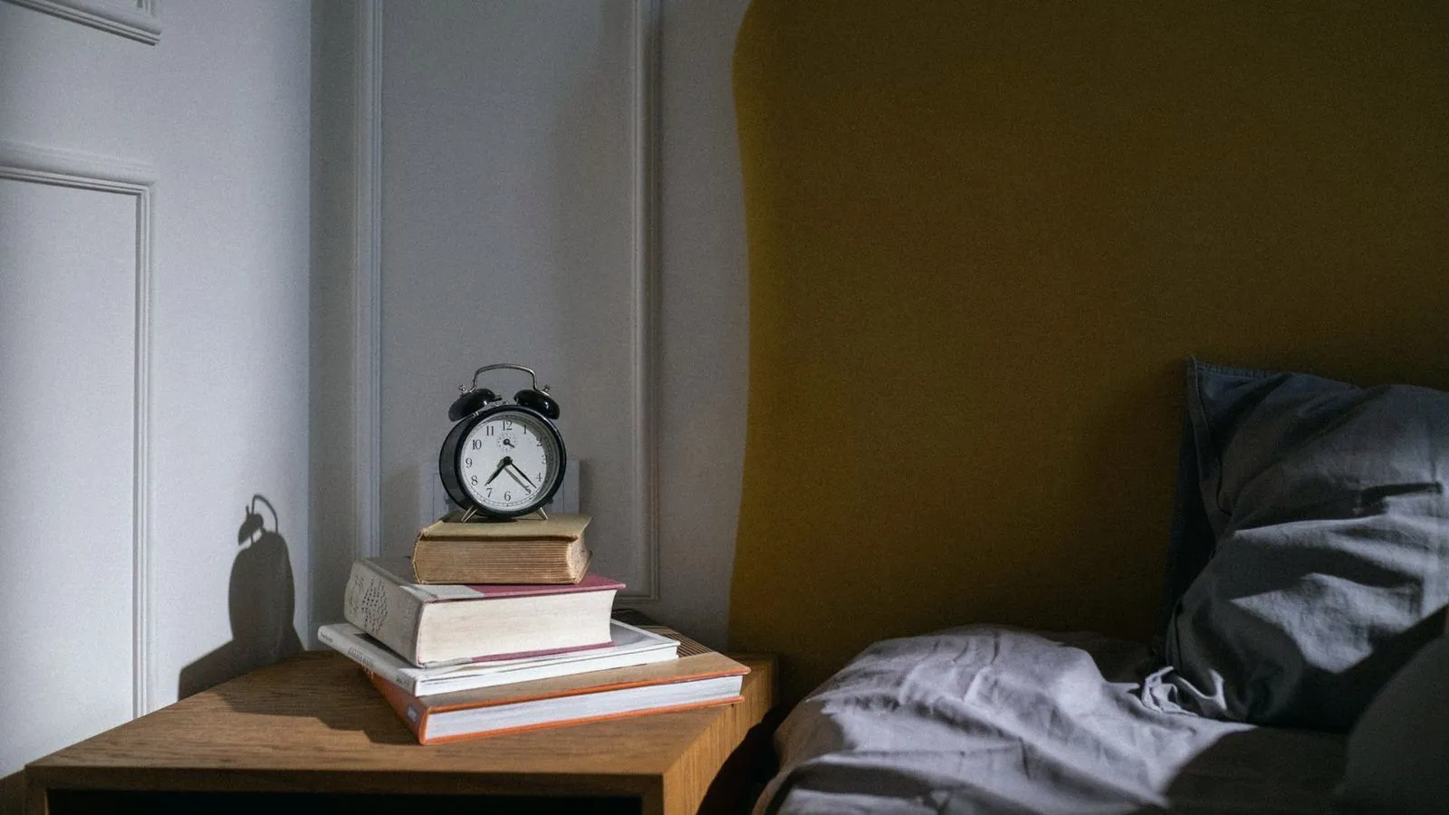 An alarm clock on a stack of books beside an unmade bed in a dim room