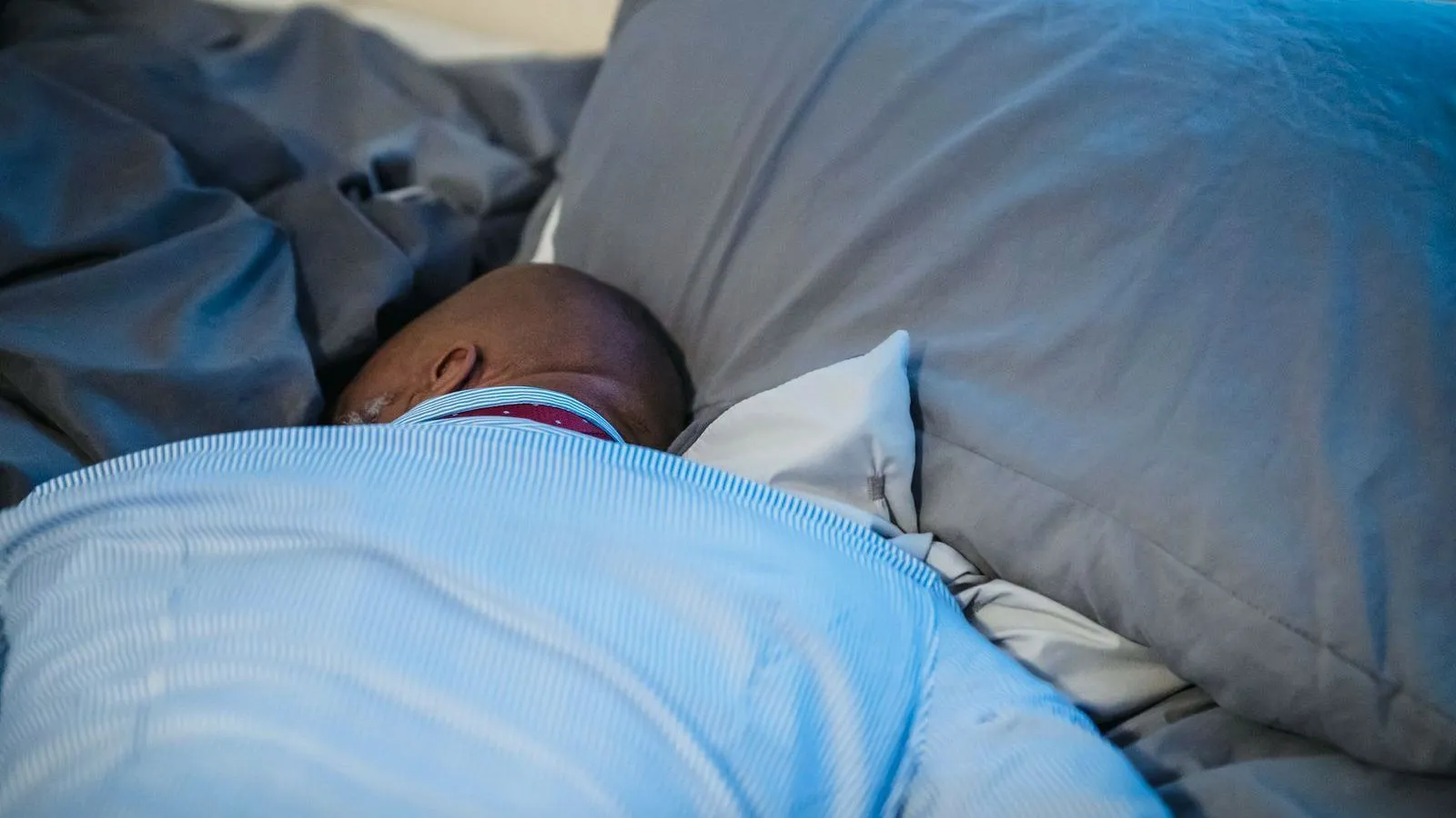 A person asleep under a blue duvet with only the top of the head showing against the pillows.