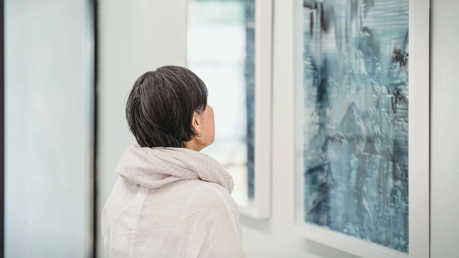 A person with short dark hair, seen from behind, looking at an abstract blue painting on a white gallery wall.