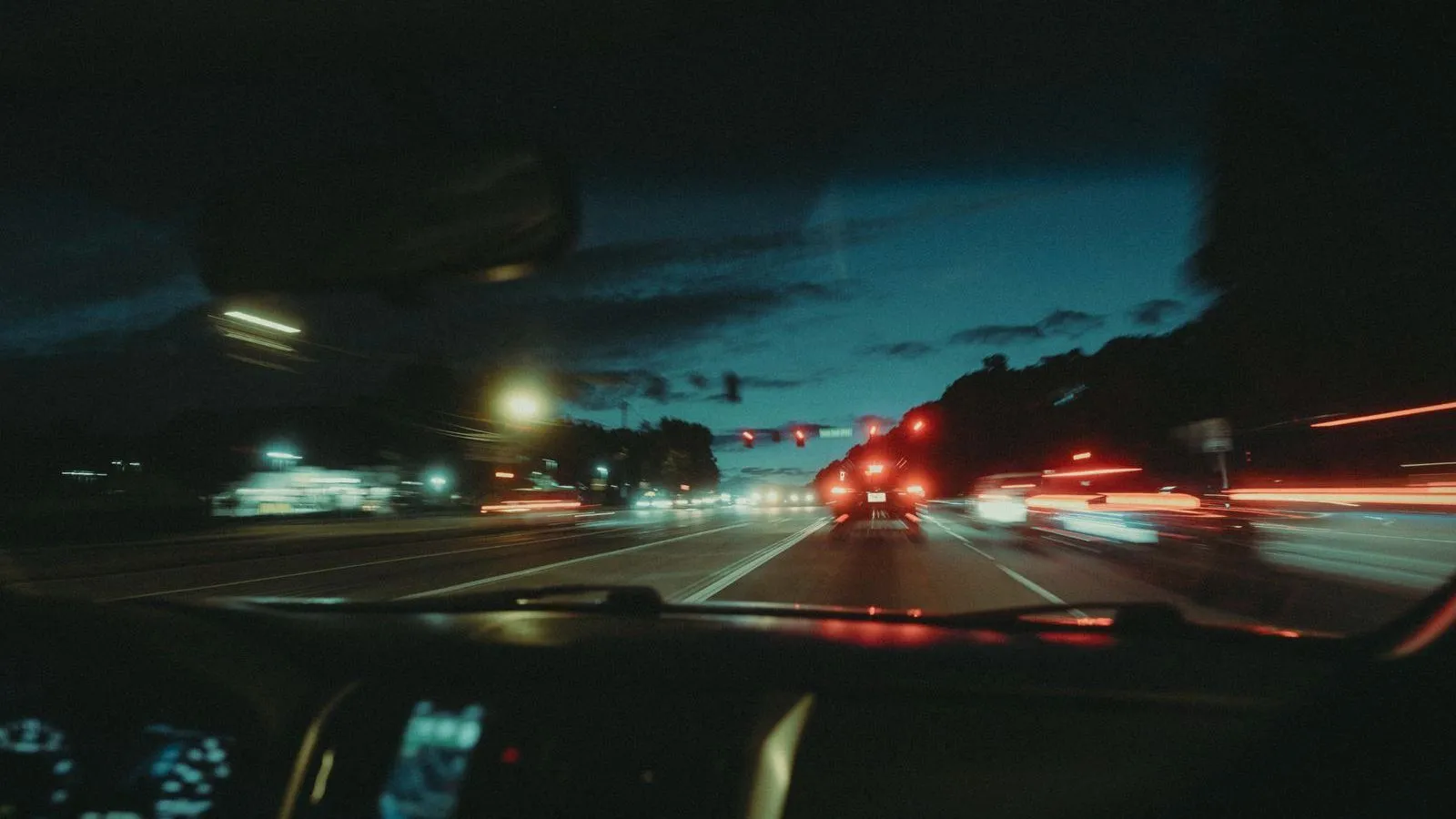 The view through a windshield of a multi-lane road at dusk, with red tail lights streaking ahead