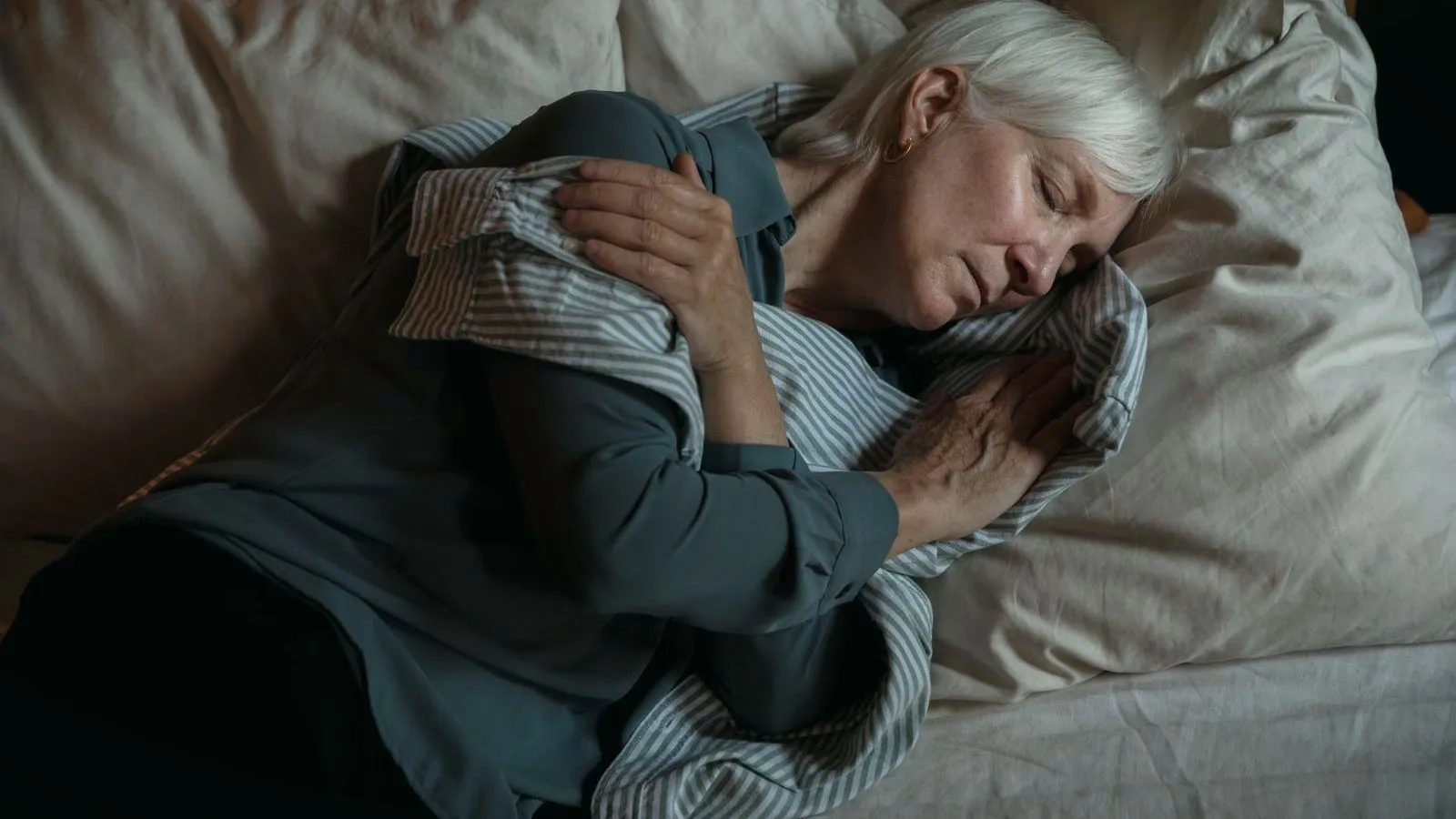 An older woman with white hair asleep on a bed, hugging a striped pillow.