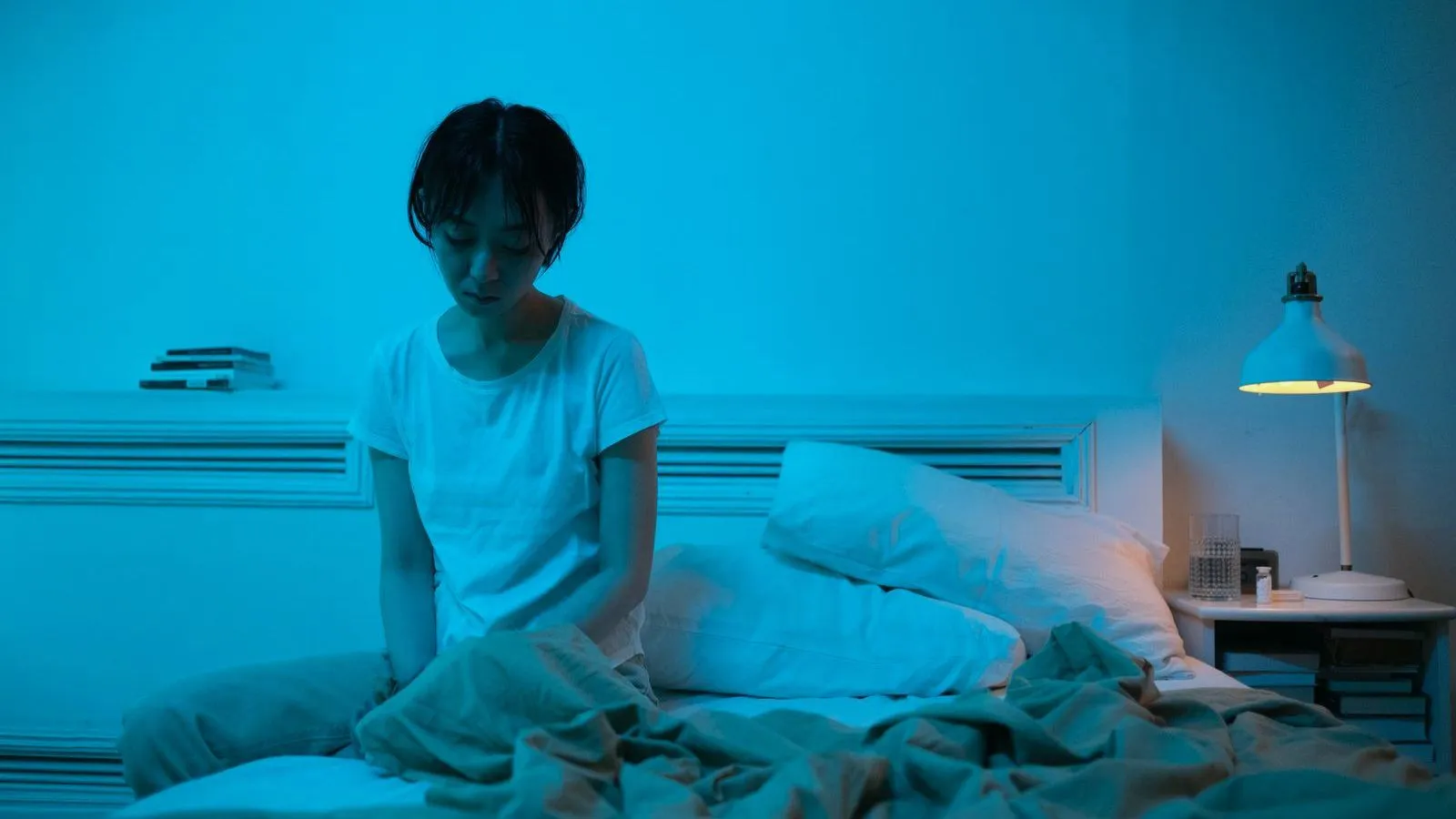 A woman sitting up awake in bed at night under blue light, a lamp glowing on the nightstand