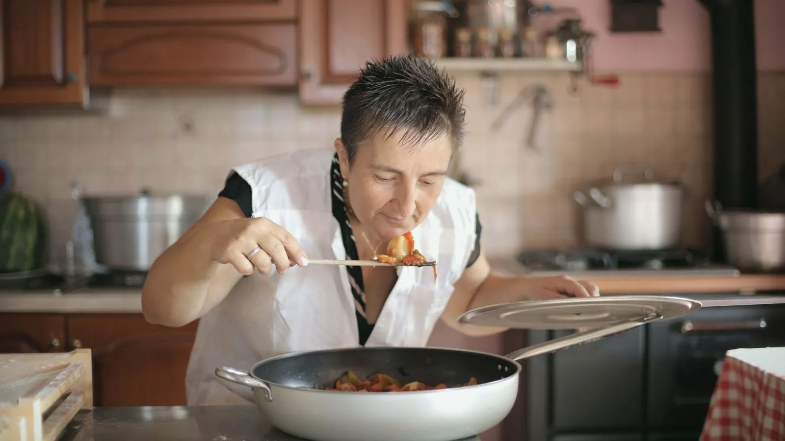 A woman in a kitchen leaning over a pan to smell food on a wooden spoon