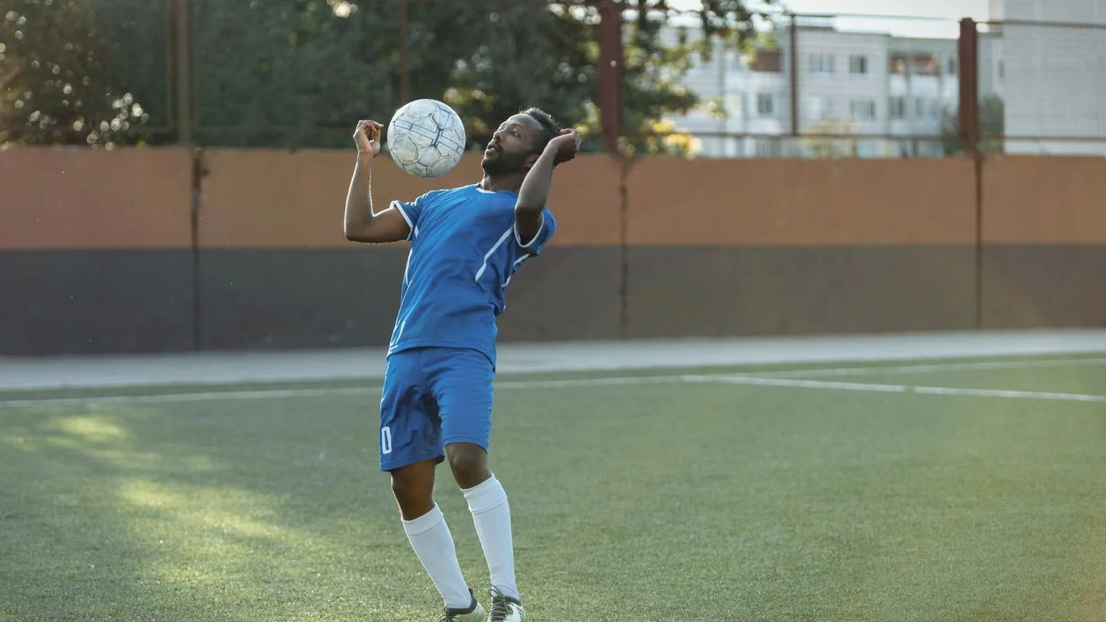 A soccer player in a blue kit and white socks leaning back with the ball at head height on an artificial pitch.