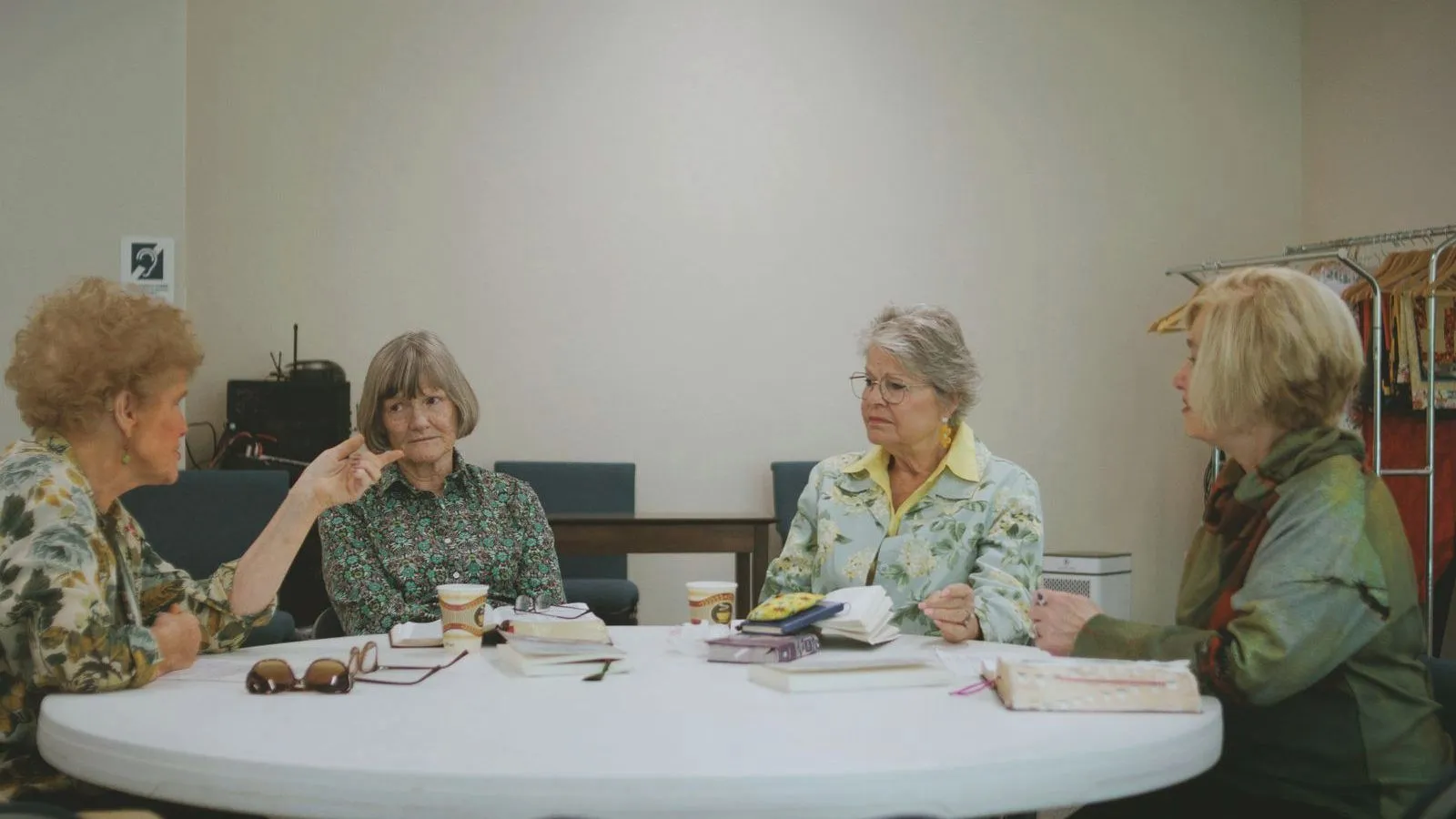Four older women sitting round a white table with books, coffee cups and reading glasses, mid-conversation