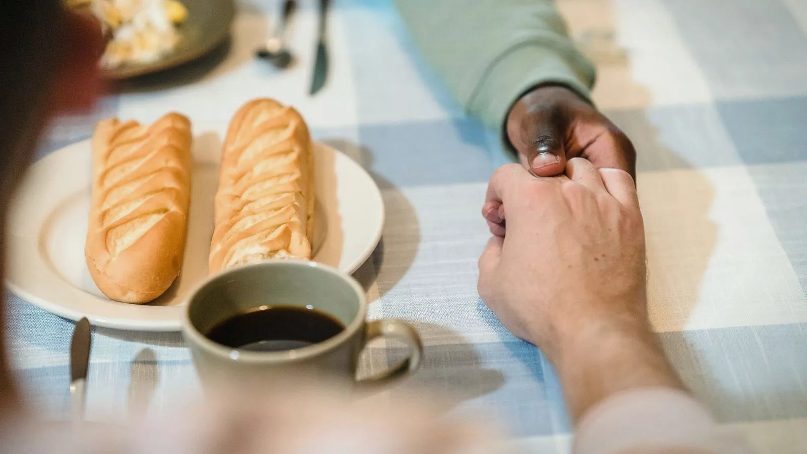 Two people holding hands across a table set with bread rolls on a plate and a cup of coffee.