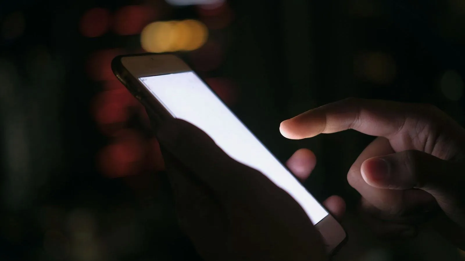 Close-up of two hands holding and tapping a smartphone with a blank glowing screen in the dark, blurred lights behind.