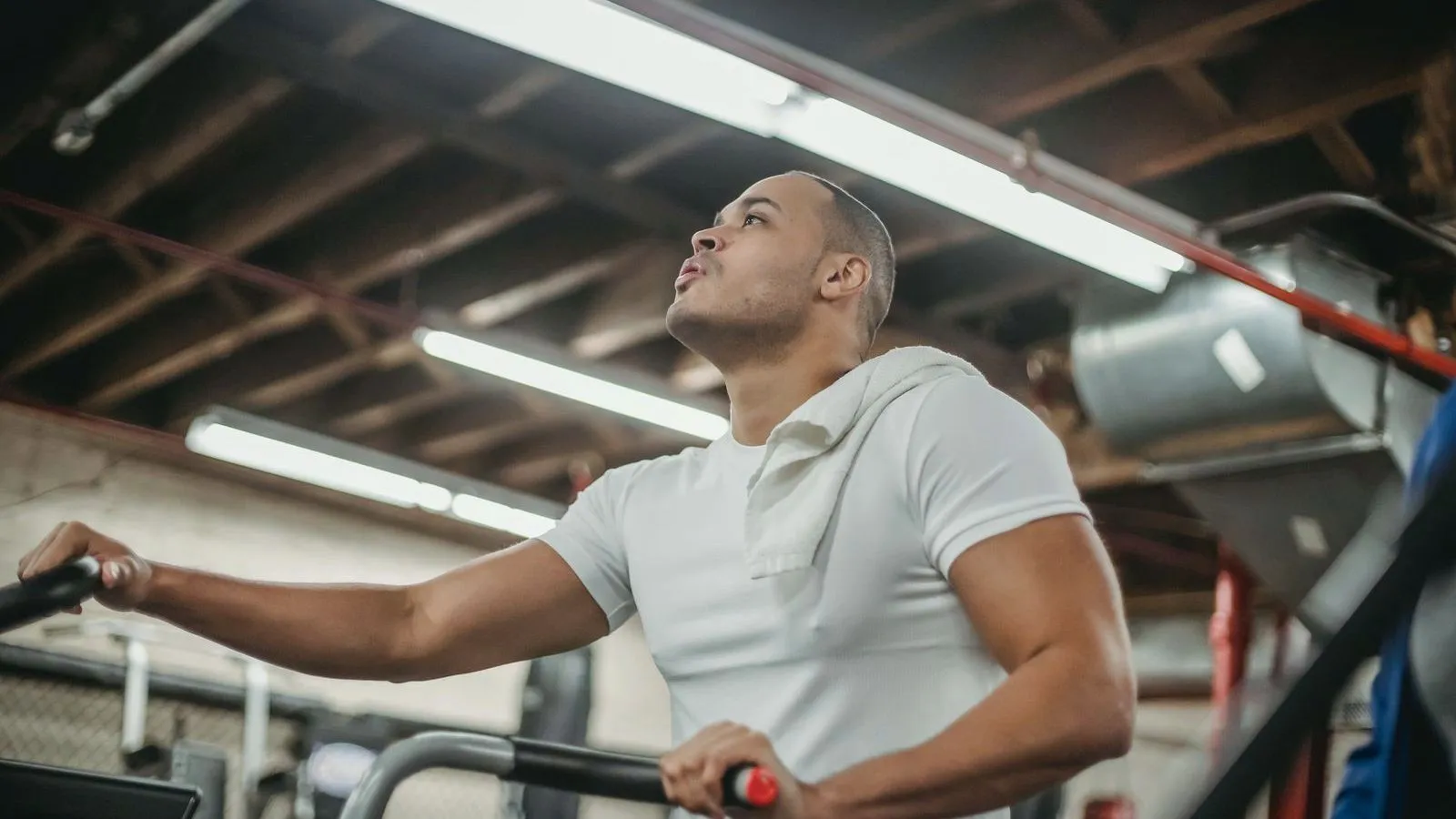 A person in a white T-shirt with a towel over one shoulder, breathing hard while gripping the handles of a gym machine.