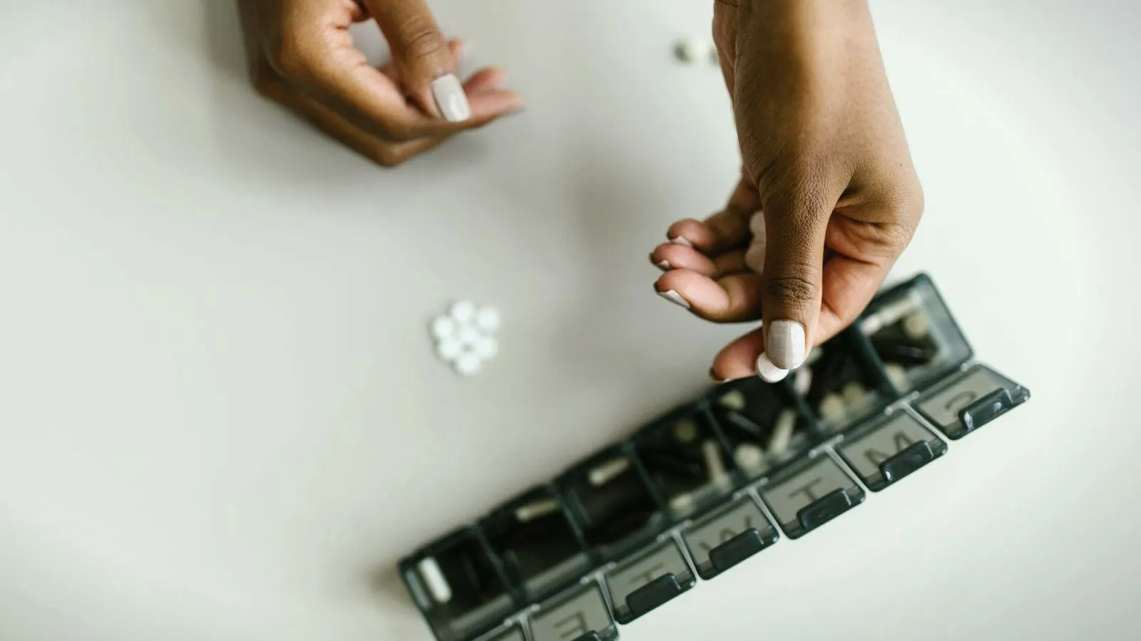 Hands placing tablets into a weekly pill organizer on a white table.