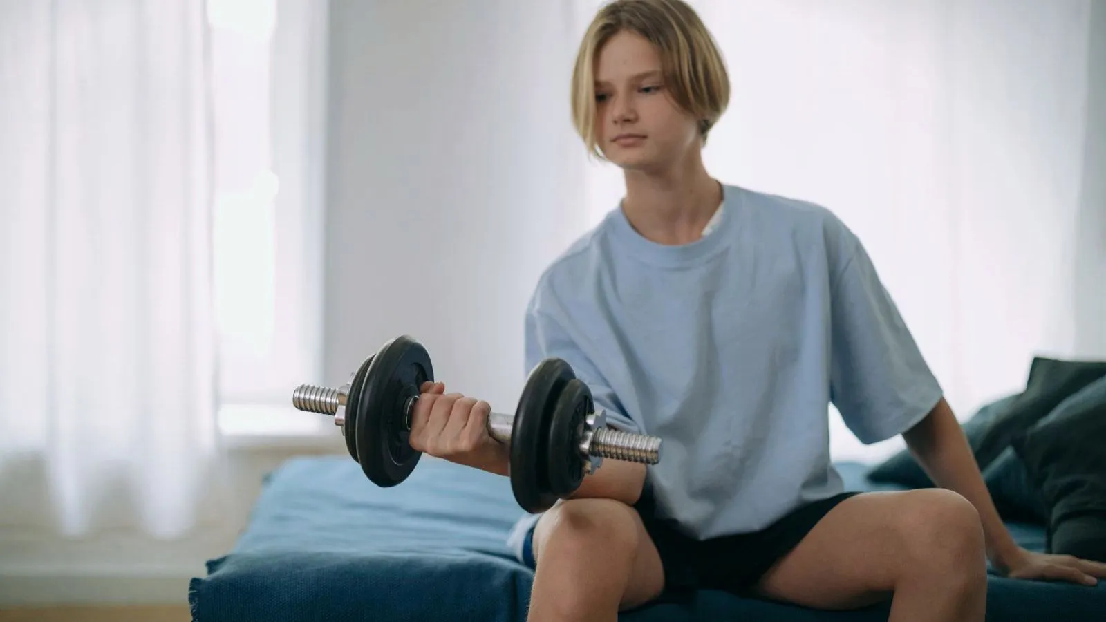 A teenager sitting on a bed curling a dumbbell.