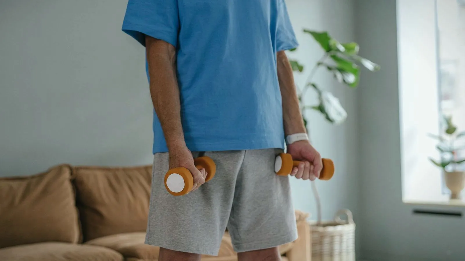 A person in a blue T-shirt and gray shorts standing in a living room holding a small orange dumbbell in each hand, face not shown.