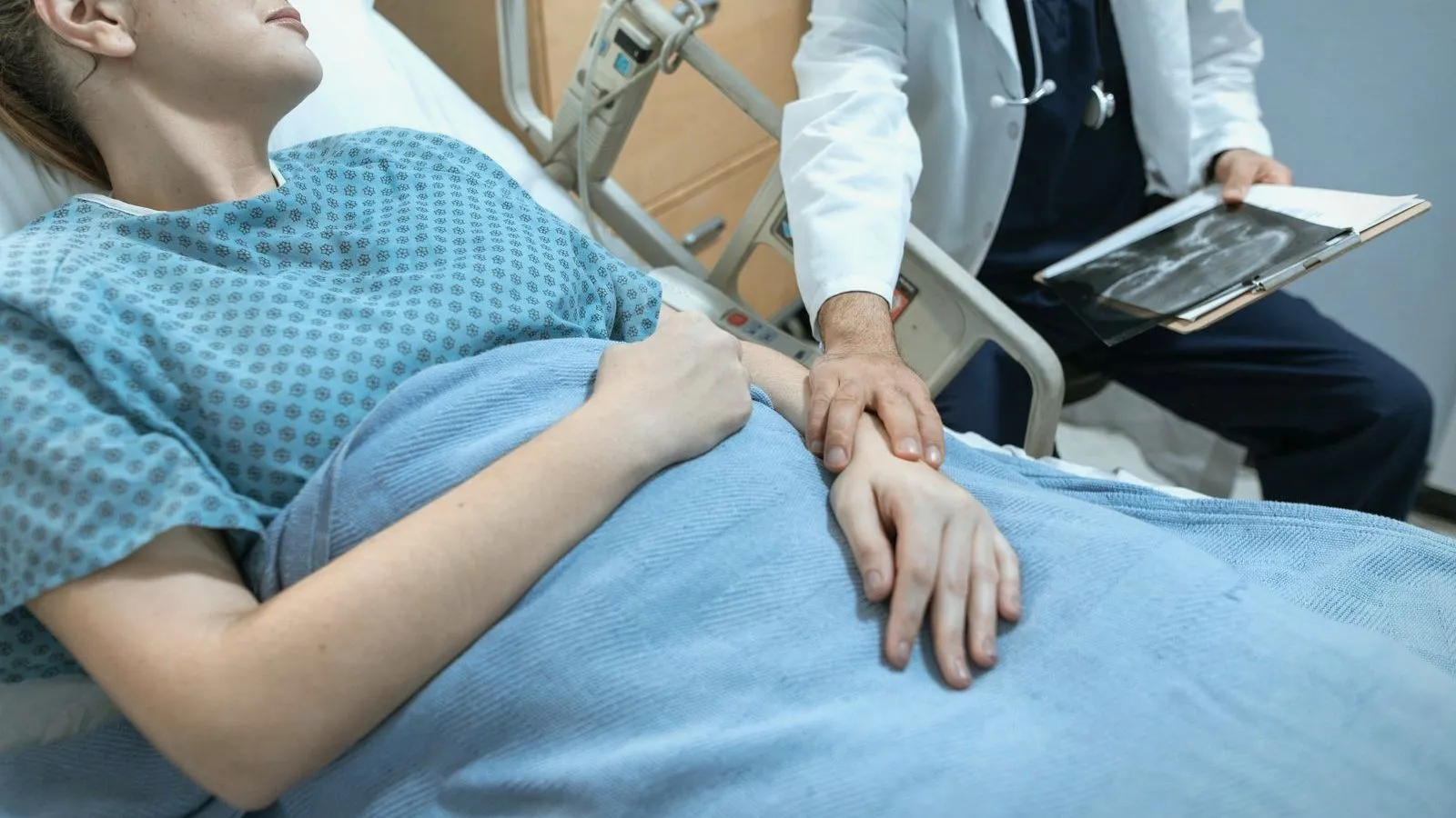 A person in a patterned hospital gown lies in a bed while a doctor in a white coat rests a hand on the person's arm and holds a scan image.