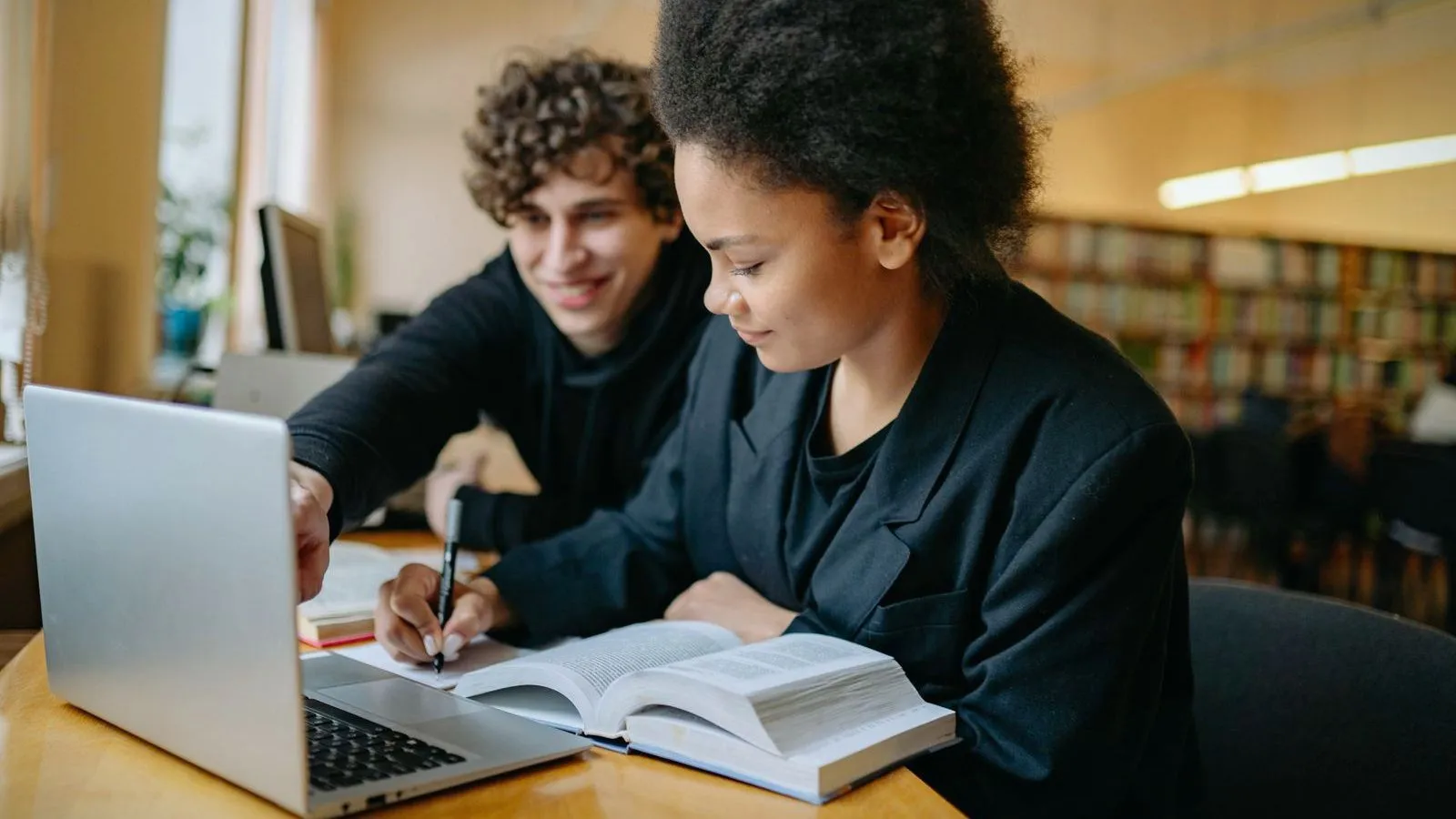 Two students working together at a library desk with a laptop and an open textbook
