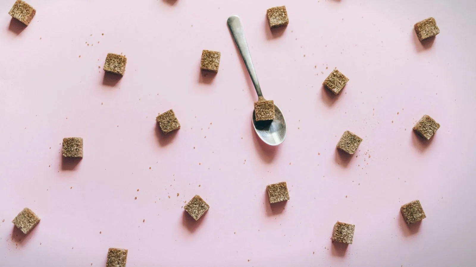 Brown sugar cubes scattered around a spoon on a pink background.
