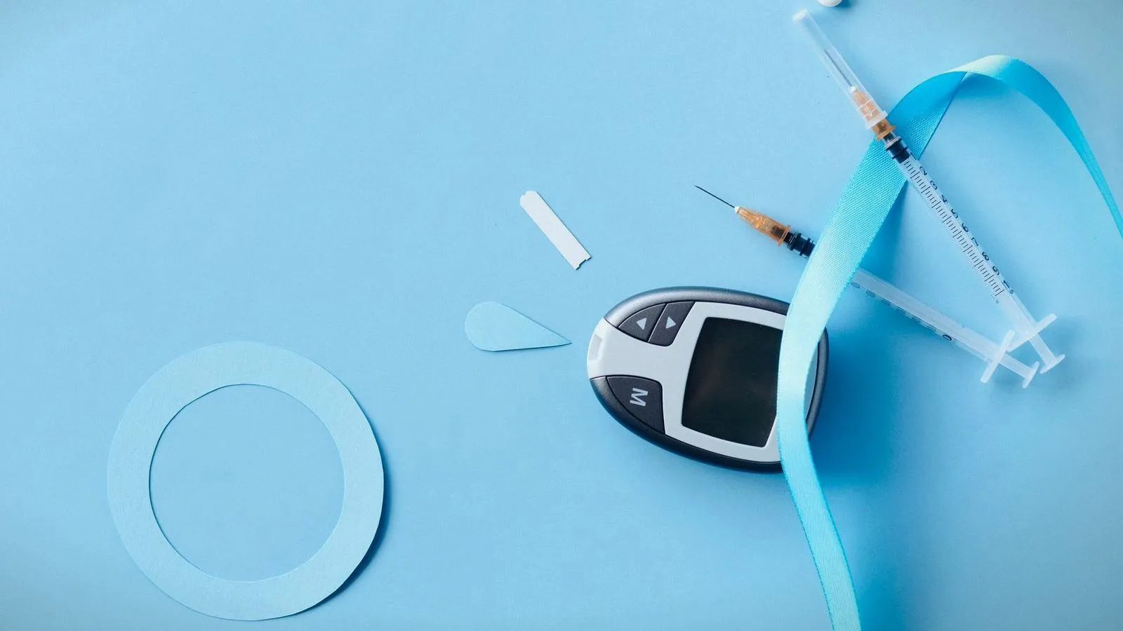 A blood glucose meter, two syringes, a test strip and a blue ribbon laid out on a pale blue surface.