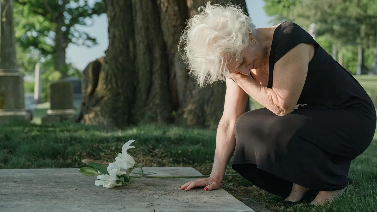 An older person in black crouching at a graveside with a hand to their face, white lilies on the stone.
