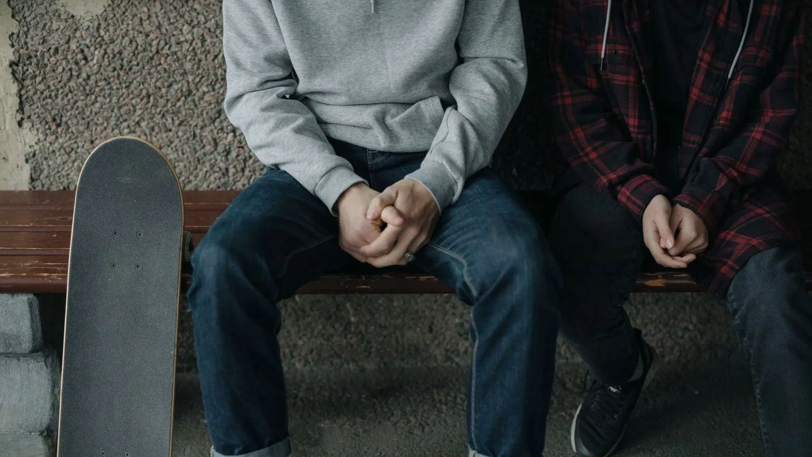 Two young people in a grey hoodie and a plaid shirt sit on a bench with hands clasped, a skateboard propped beside them.